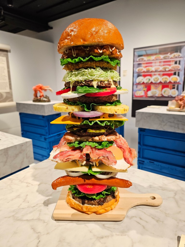An absurdly tall burger with an abundance of ingredients, including patties, vegetables, cheese and bacon. The burger is a food replica.