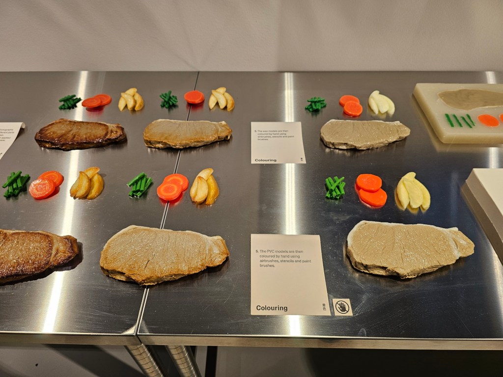 A middle stage of creating a food replica: steak with carrots, beans and potatoes, served on a plate. The silicone models are being painted.