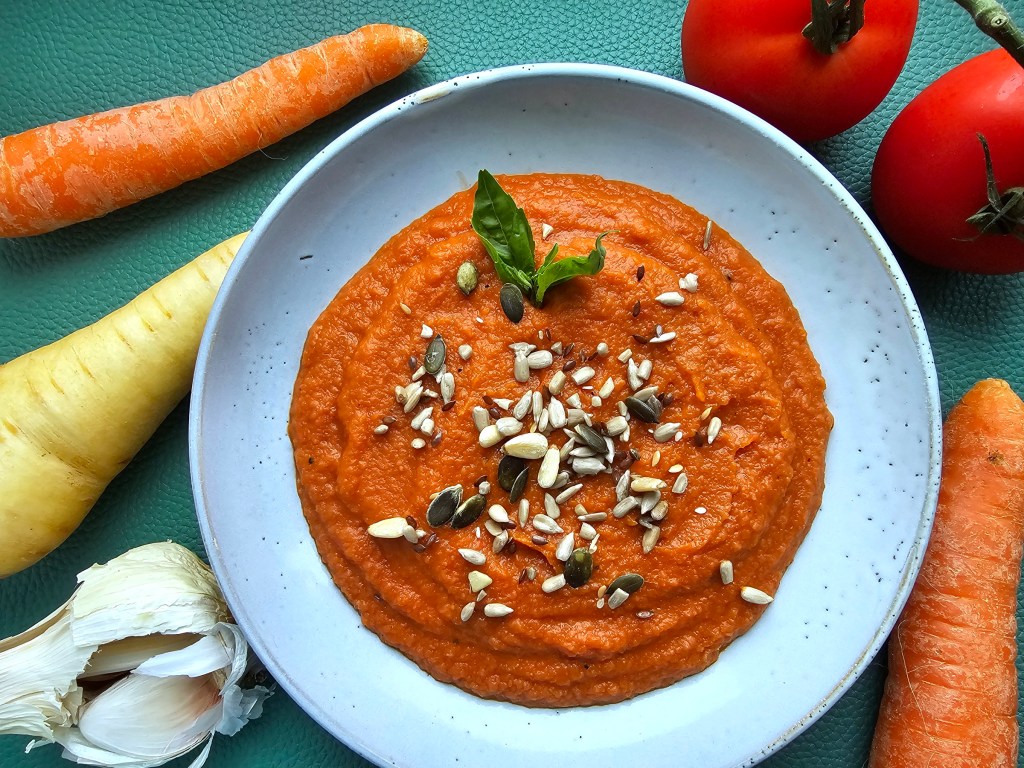 Creamy tomato and roasted vegetable soup in a blue plate, garnished with mixed seeds and fresh basil.