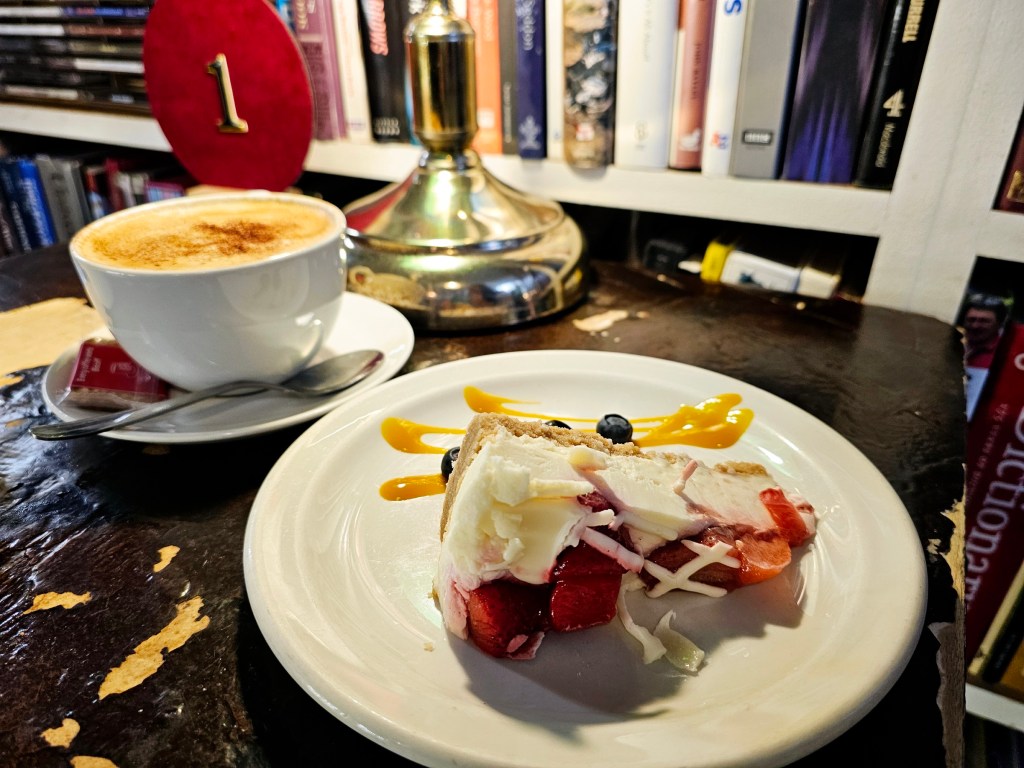 A large, white cup with a cappuccino and a slice of strawberry cheesecake on a plate.