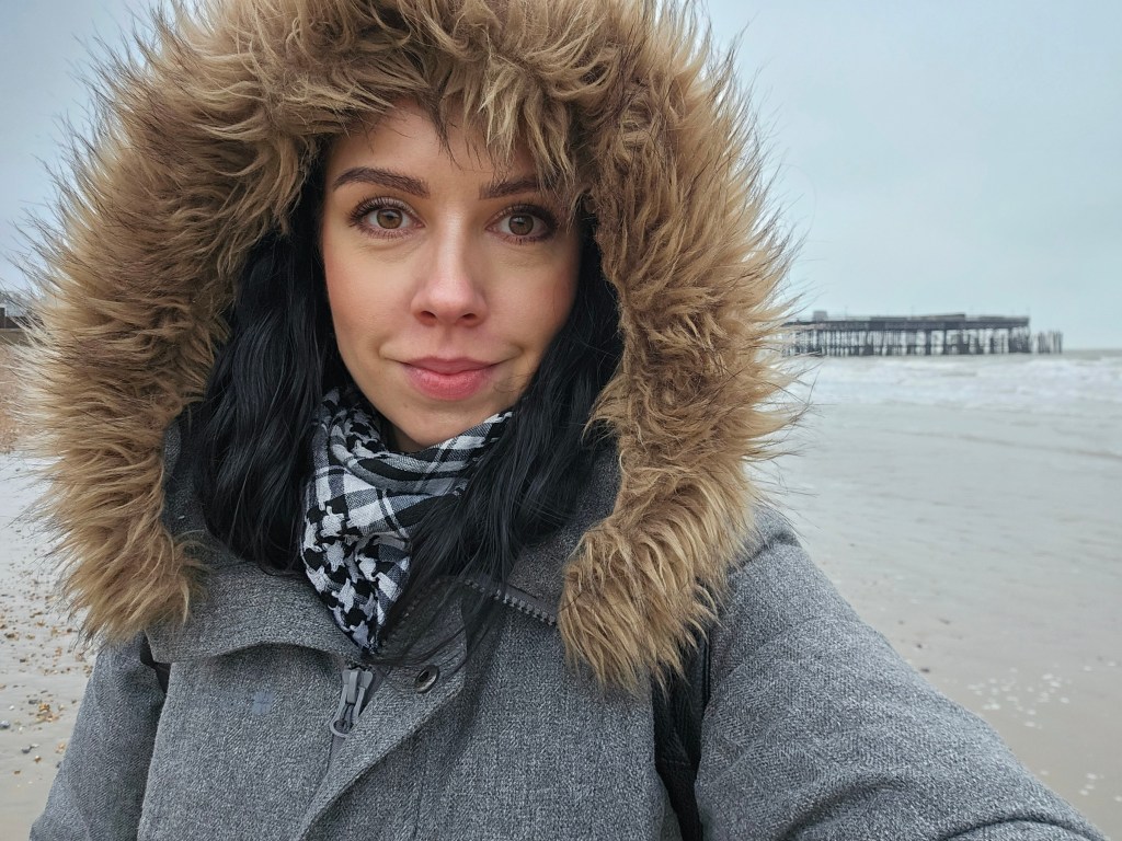A selfie on the Hastings beach. The woman is wearing a warm winter coat with a furry hood.