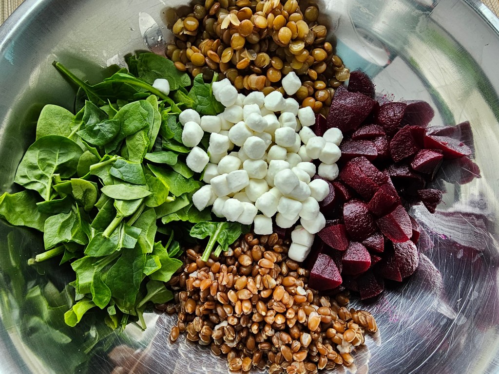 Baby spinach, cooked green lentils and wheatberries, goat's cheese and pickled beetroot in a metal bowl.