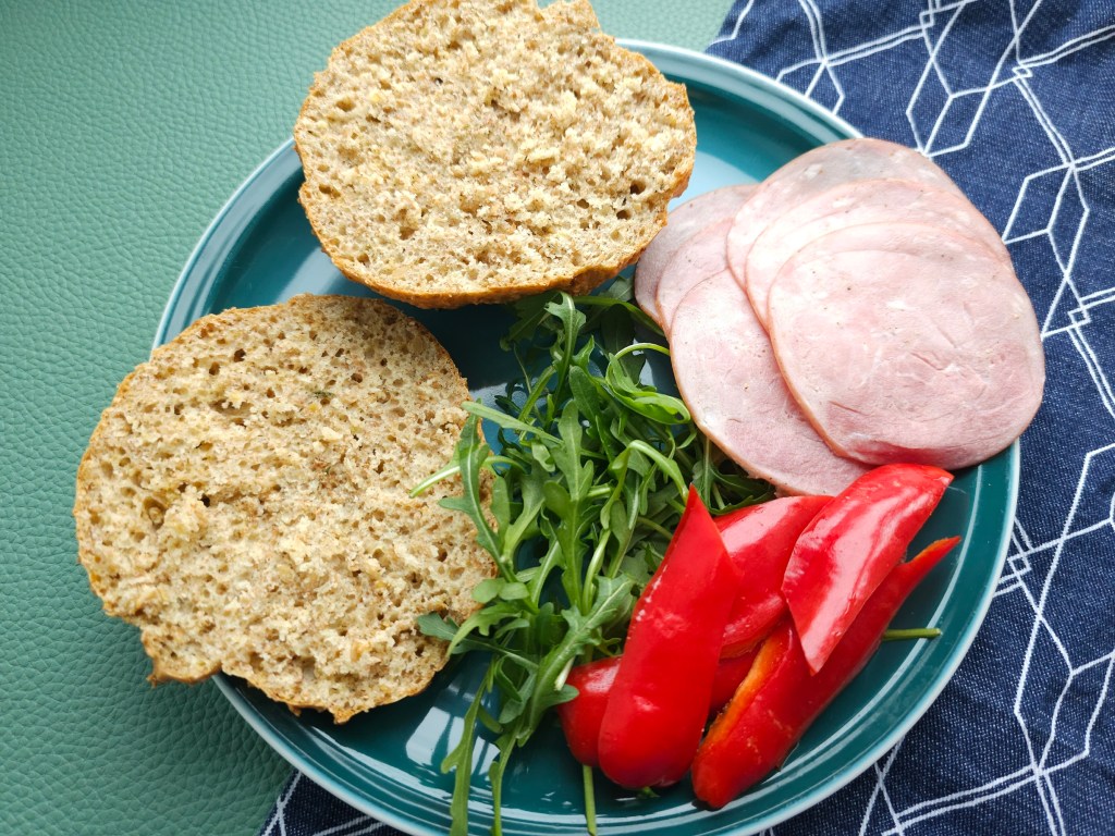 A homemade cottage cheese bread roll sliced in half, on a plate, next to rocket, sliced red bell pepper and a few slices of ham.
