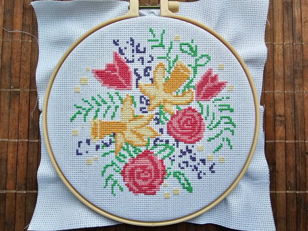 Nearly finished cross stitch design featuring daffodils, roses and tulips. Only the leaves and purple flowers are missing details.
