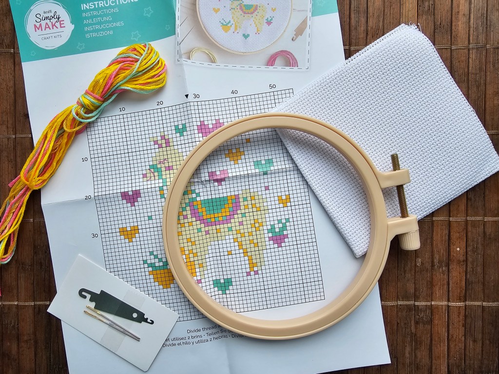 Contents of a mini DIY cross stitch kit featuring a llama design. The kit includes a blank aida, a plastic hoop, a couple of needles, a threader and yarn in assorted colours.