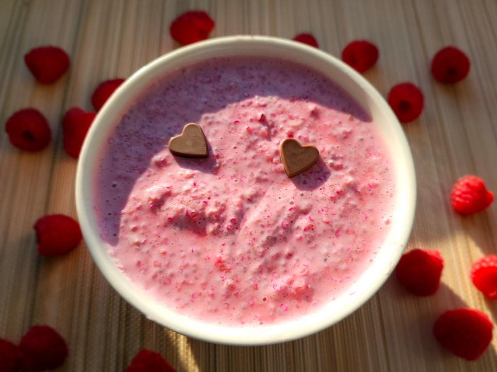A bowl with raspberry quinoa chia pudding. Fresh raspberries scattered around the bowl. Two chocolate hearts are placed on top of the pudding for decoration.