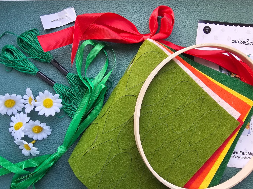 Contents of a DIY craft kit for making a wreath with felt flowers: a wooden hoop, 6 daisies, ribbon, thread and felt sheets with pre-cut petals.