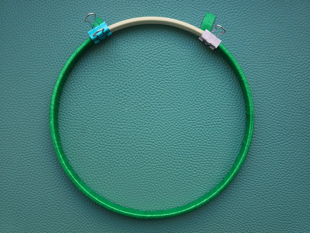 Wooden hoop wrapped with green ribbon. The ends of the ribbon are held in place by bag clips. There's not enough ribbon to cover the whole hoop.