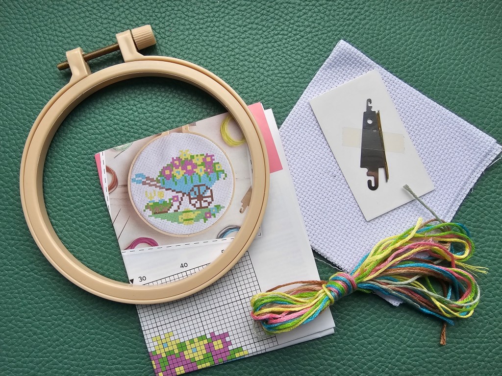 Contents of a mini cross stitch kit. It includes a blank aida, a plastic hoop, yarn in assorted colours, needles and a threader.
