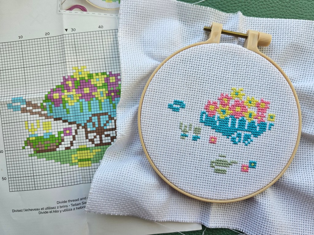 Half completed small cross stitch design featuring a wheelbarrow, gardening tools and flowers. Green and brown details are missing.