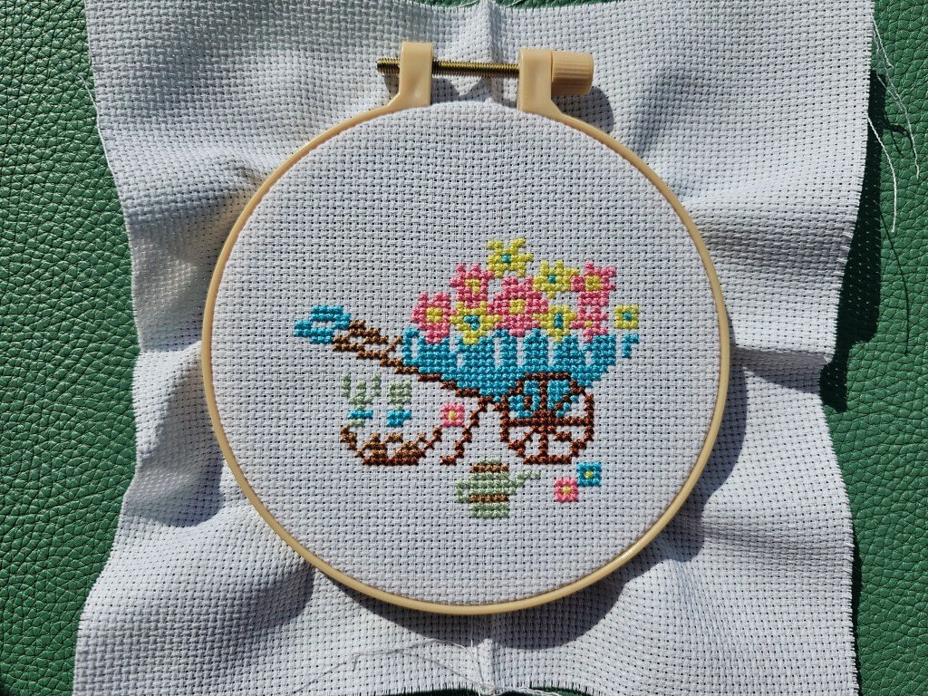 Nearly completed small cross stitch design featuring a wheelbarrow, gardening tools and flowers. Green details are missing.