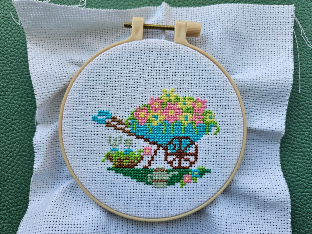 Completed small cross stitch design featuring a wheelbarrow, gardening tools and flowers.