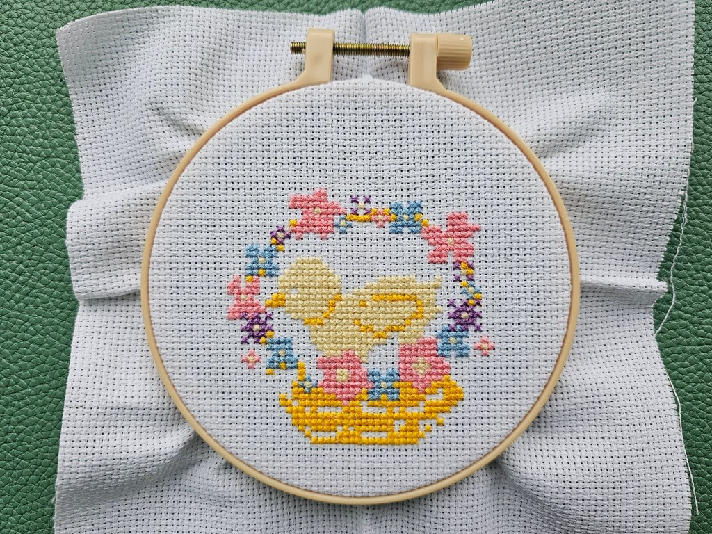 Partially completed cross stitch project. The yellow chick, pink, blue and purple flowers and the basket are done.