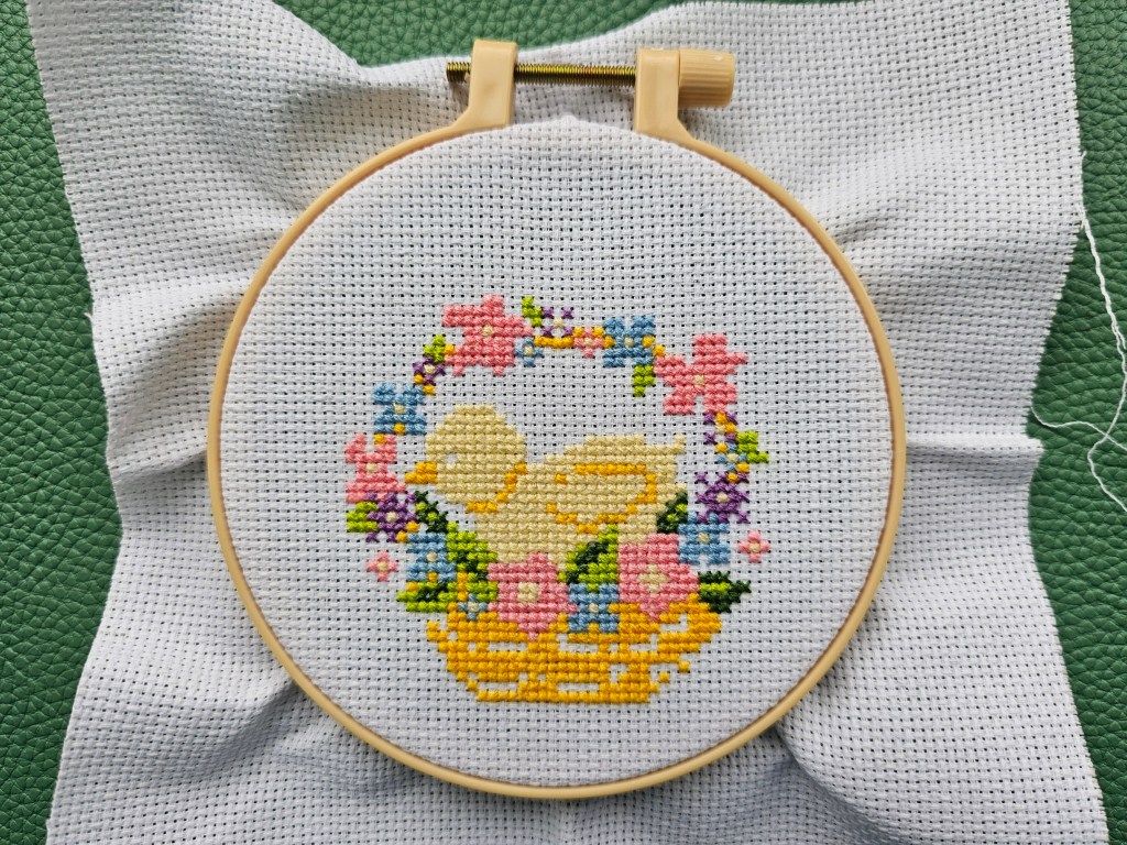 Nearly finished cross stitch design featuring a chick inside a basket decorated with flowers.