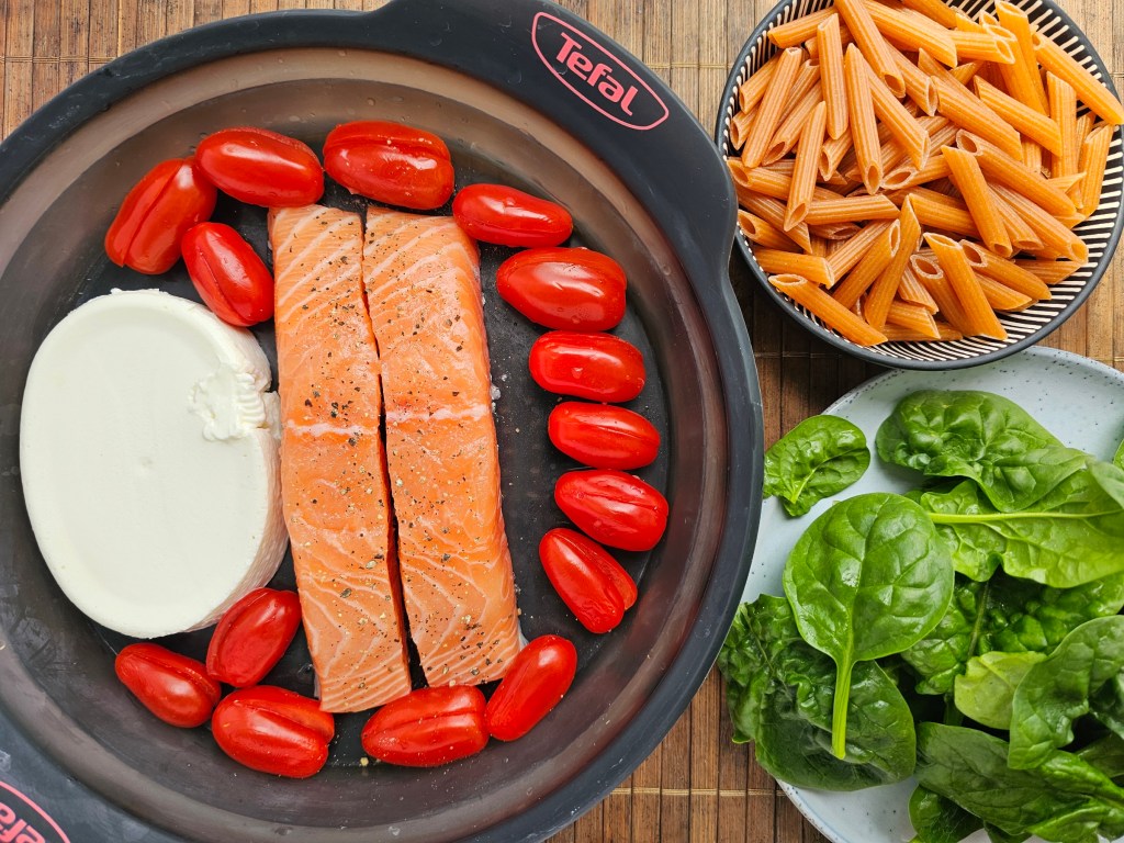 Salmon fillets, cream cheese and cherry tomatoes, before baking. A bowl of uncooked pasta and raw spinach.