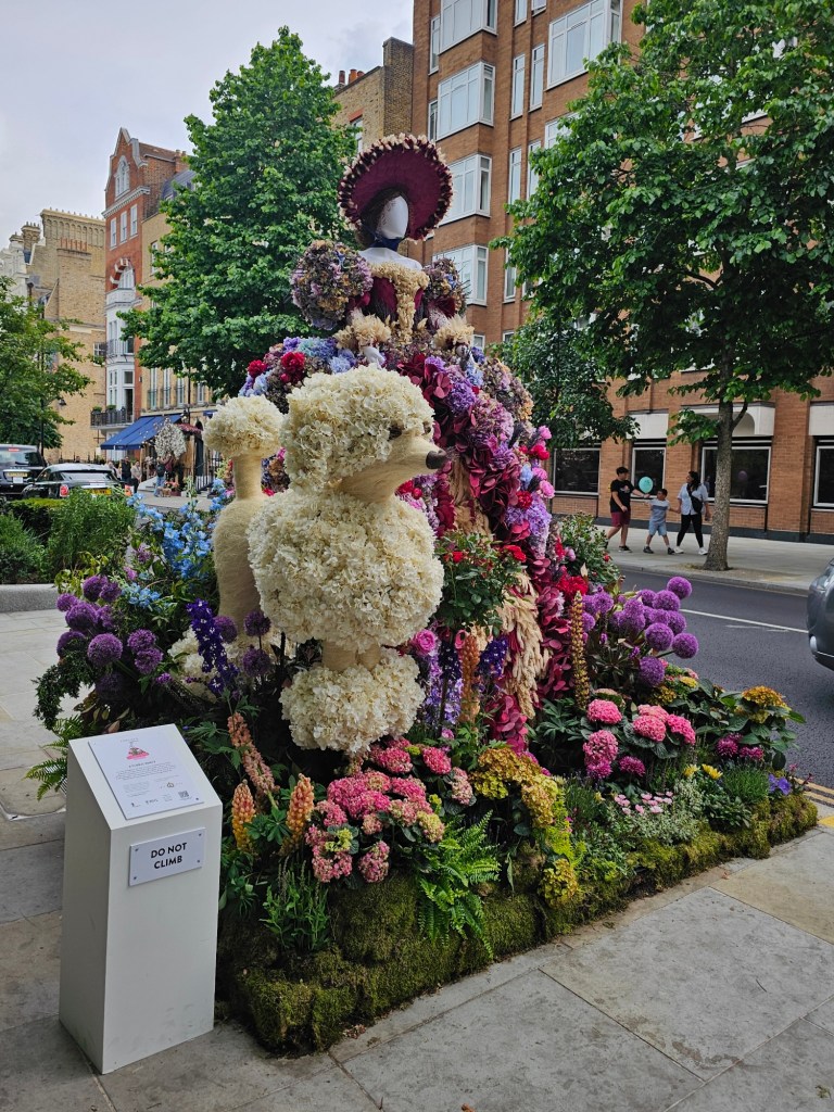 A floral display featuring a person wearing a colourful princess dress, and a white poodle dog.