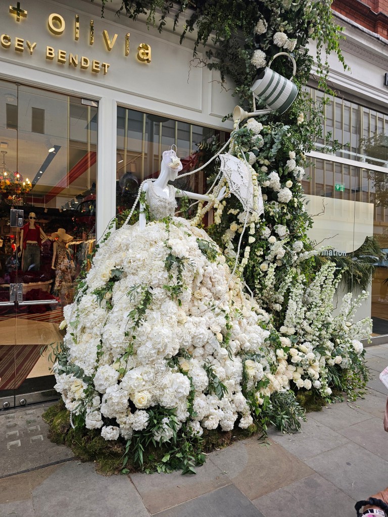 A beautiful floral display next to a shop entrance. It features a person wearing a white princess dress made of white flowers.