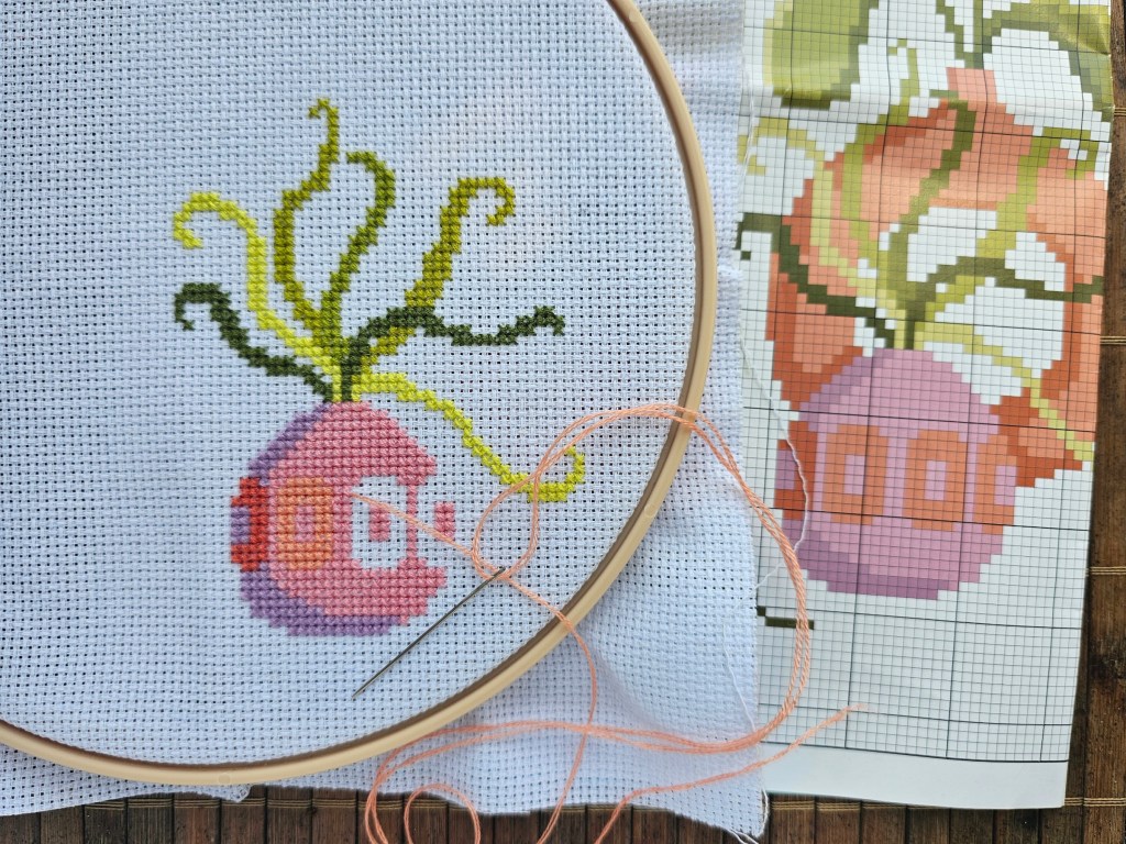 A vase with green plants cross stitched onto a blank aida.