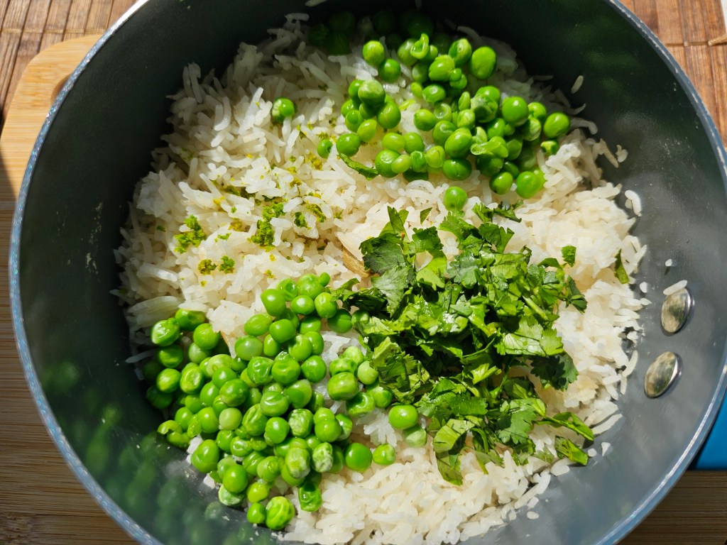 A pot with cooked white rice, chopped up coriander leaves, lime zest and green peas.