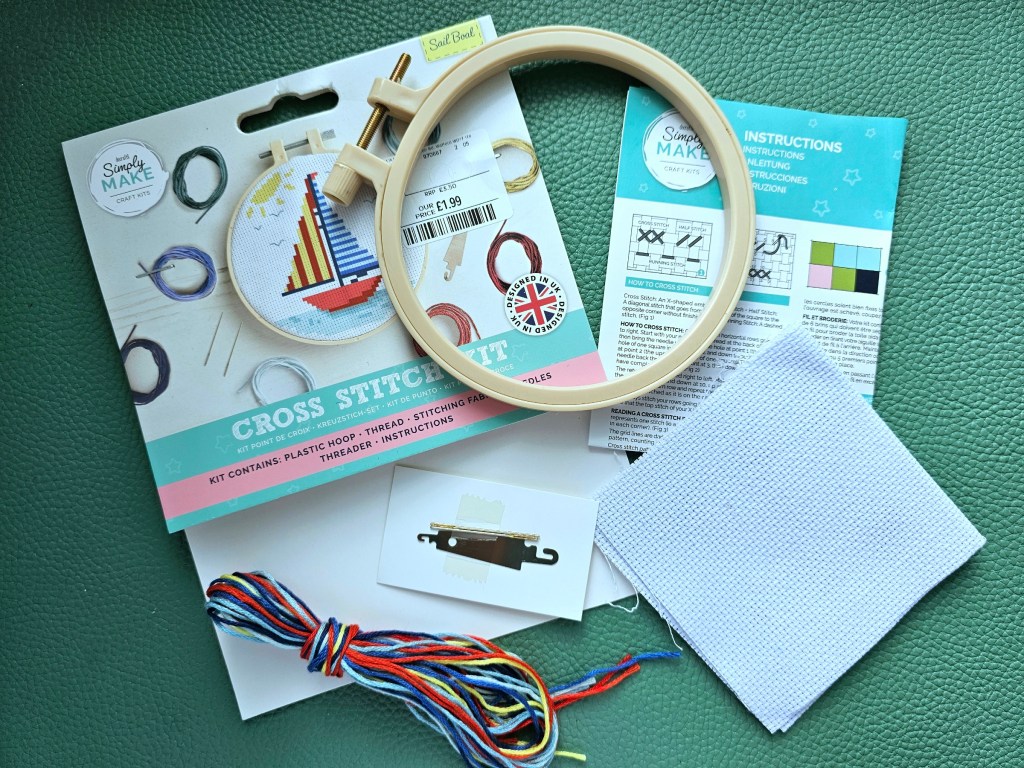 Contents of a DIY cross stitch kit with a sailboat design. Blank aida, threads in assorted colours, needles, threader and an embroidery hoop.