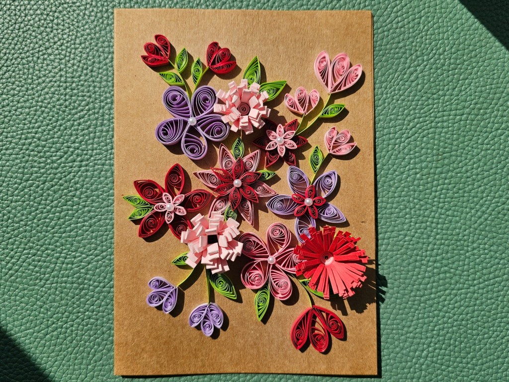 A handmade card featuring lots of pink, red and purple quilled flowers in various styles.