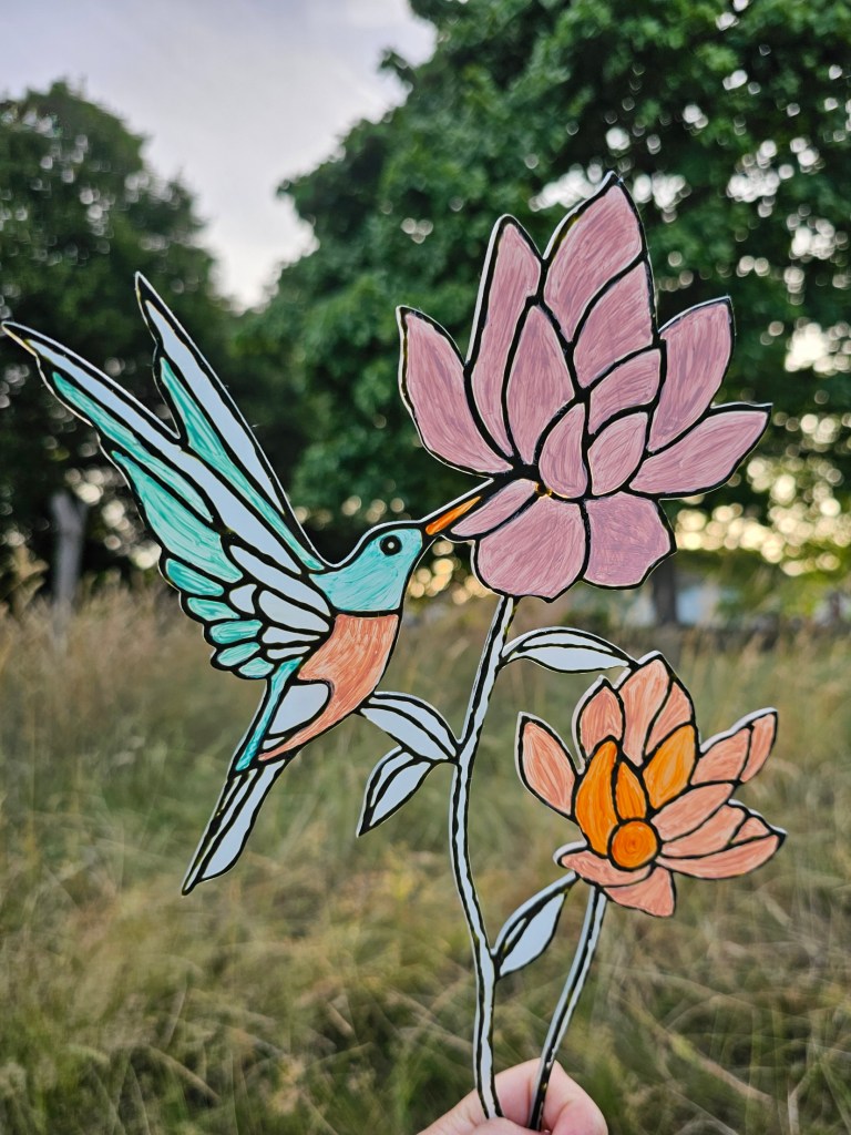 Partially finished fake stained glass ornament. Two flowers are painted with pink acrylic paint, and the hummingbird is green.