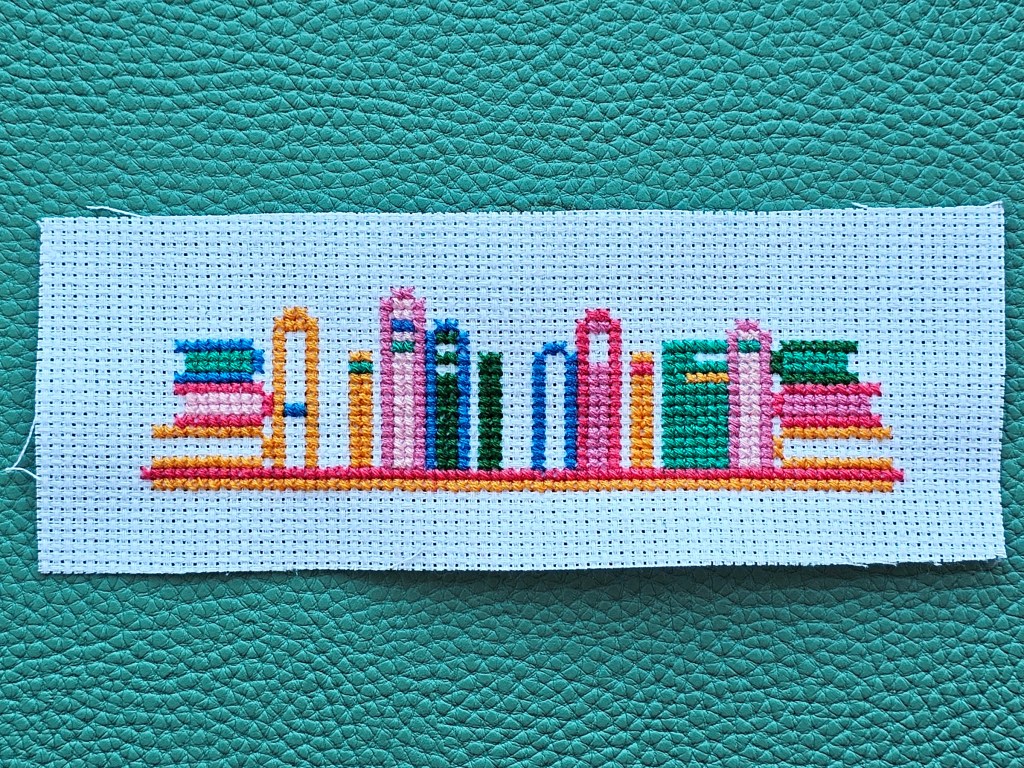 Nearly completed cross stitch bookmark with a design featuring a shelf with colourful books.