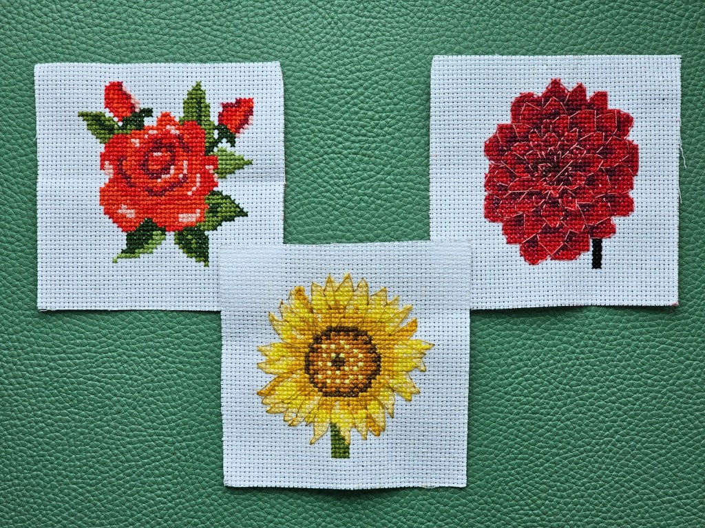 Three completed botanical mini cross stitch designs, featuring a rose, a sunflower and a dahlia