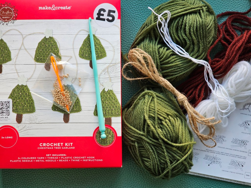 Contents of a crochet kit for making a garland of Christmas trees - it includes yarn in a few colours, sewing thread, a plastic crochet hook, beads, a plastic tapestry needle and a leaflet with the pattern.