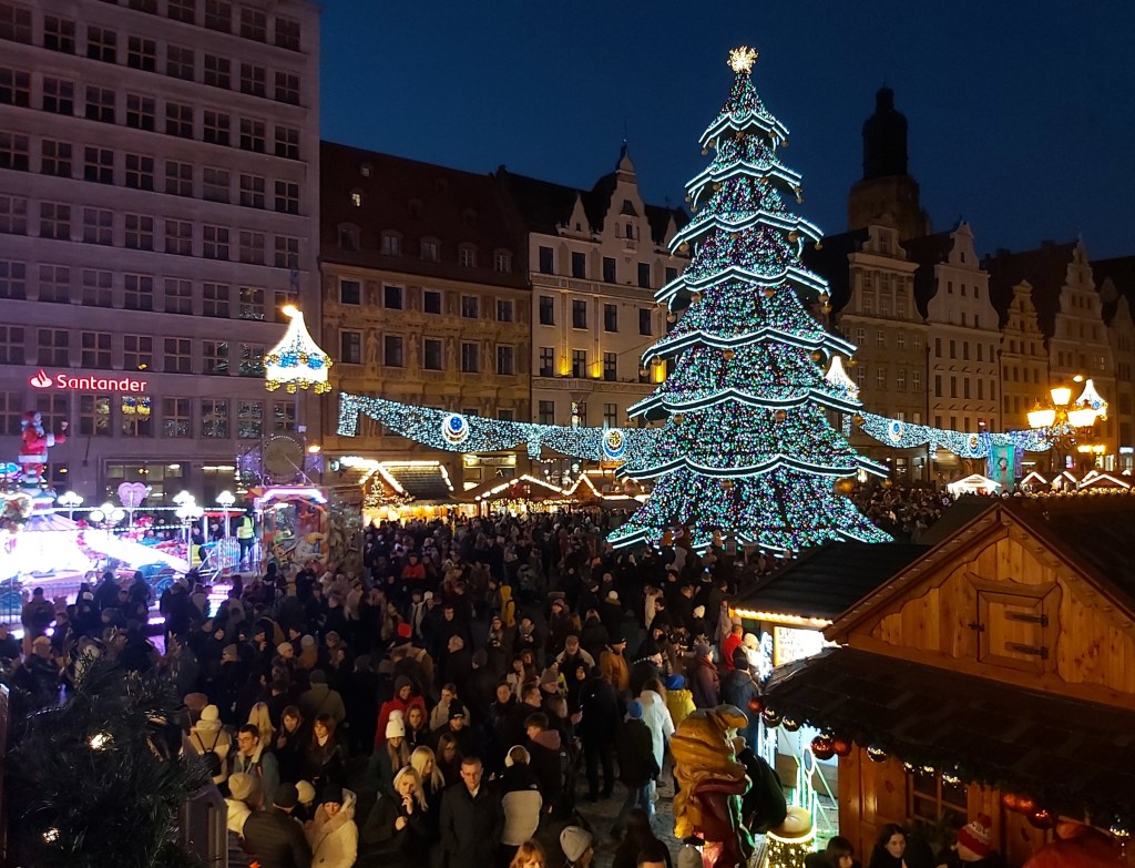 Wroclaw Christmas market by night. There are various stands, crowds of people, and a huge Christmas tree