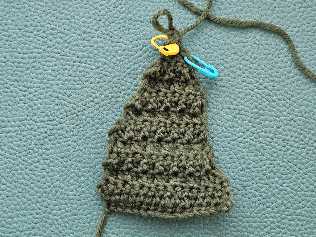 Crocheting a Christmas tree - a slightly wonky green triangle with uneven edges. Stitch markers are still attached.
