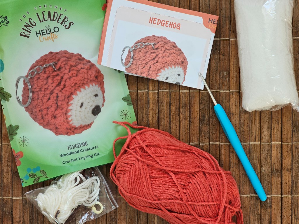 Contents of a crochet set for making a mini hedgehog keychain. There is yarn in a few colours, a metal crochet hook, toy stuffing, an empty keyring, and a leaflet with the pattern.