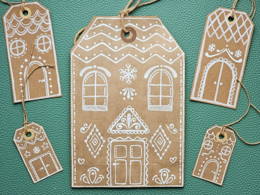 A brown paper gift bag with a gingerbread house drawn with white pen. Next to it, there are gift tags with small gingerbread houses drawn.