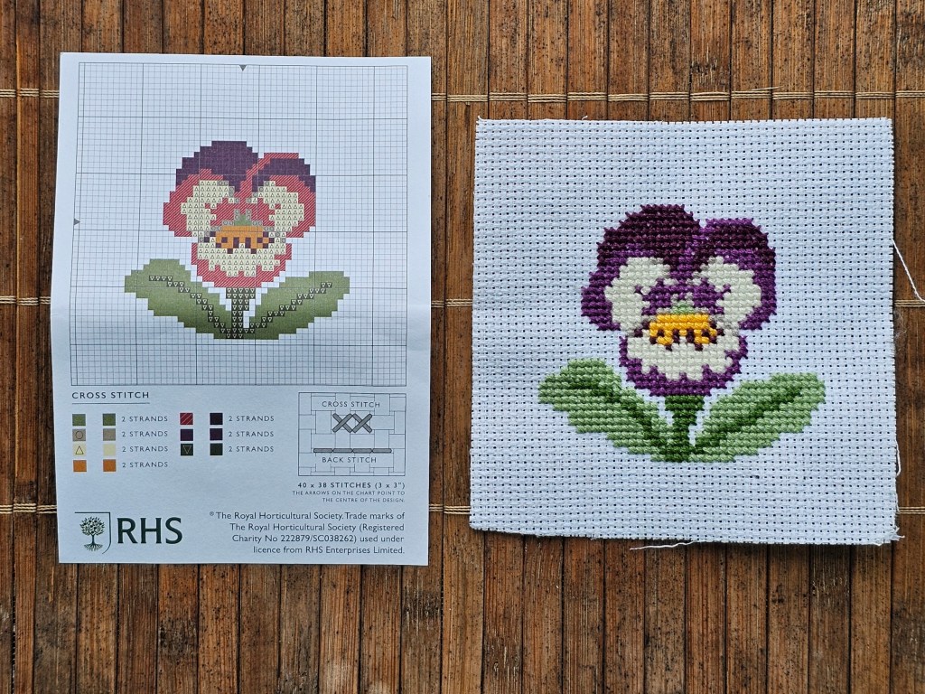 Completed cross stitch pansy next to a leaflet with the pattern