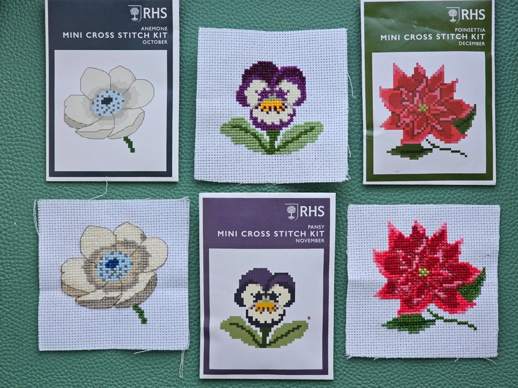 Three completed mini botanical cross stitch projects: an anemone, pansy and poinsettia