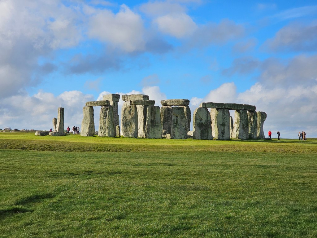 Stonehenge seen on a sunny day. The grass is green and the sky is blue. A few people can be seen next to the Stone Circle for scale.