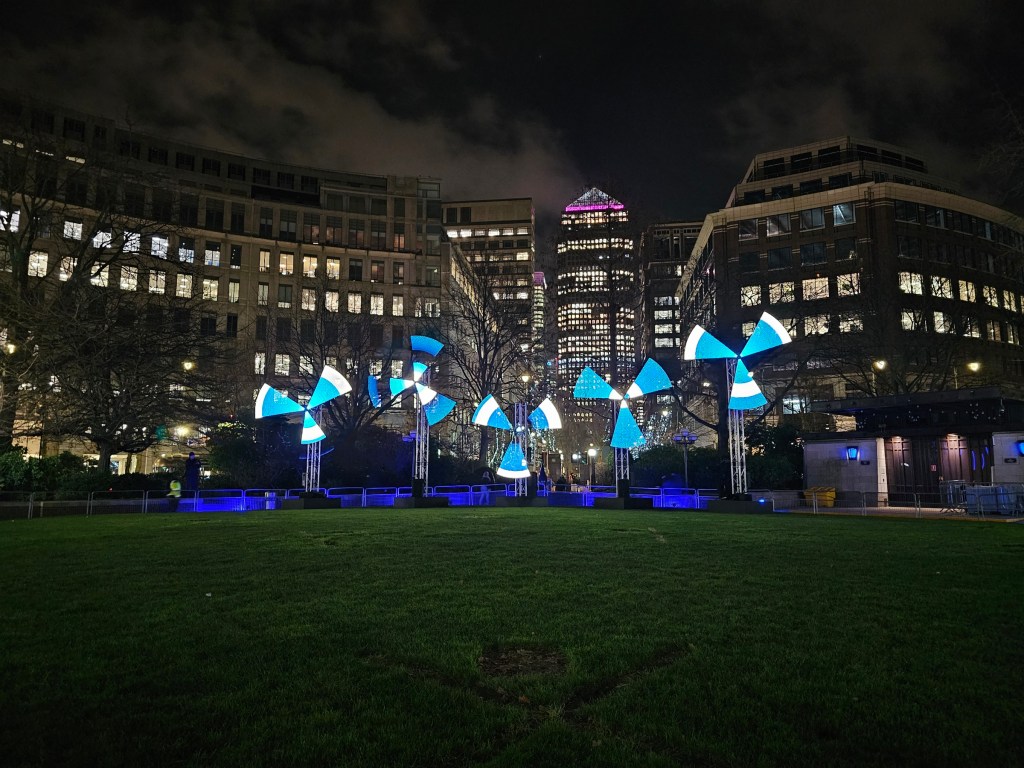 Five installations resembling rotating fans made of light. Photo taken at night in an urban area during a festival.