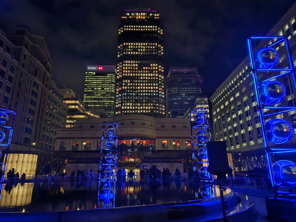 Blue light installations in Canary Wharf - four blue pillars standing on the outside of a fountain