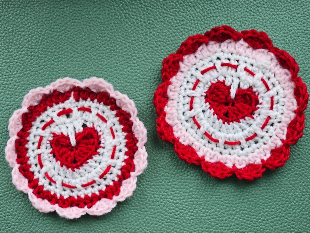 Two completed crocheted coasters. They are white, pink and red, and have a heart in the middle.