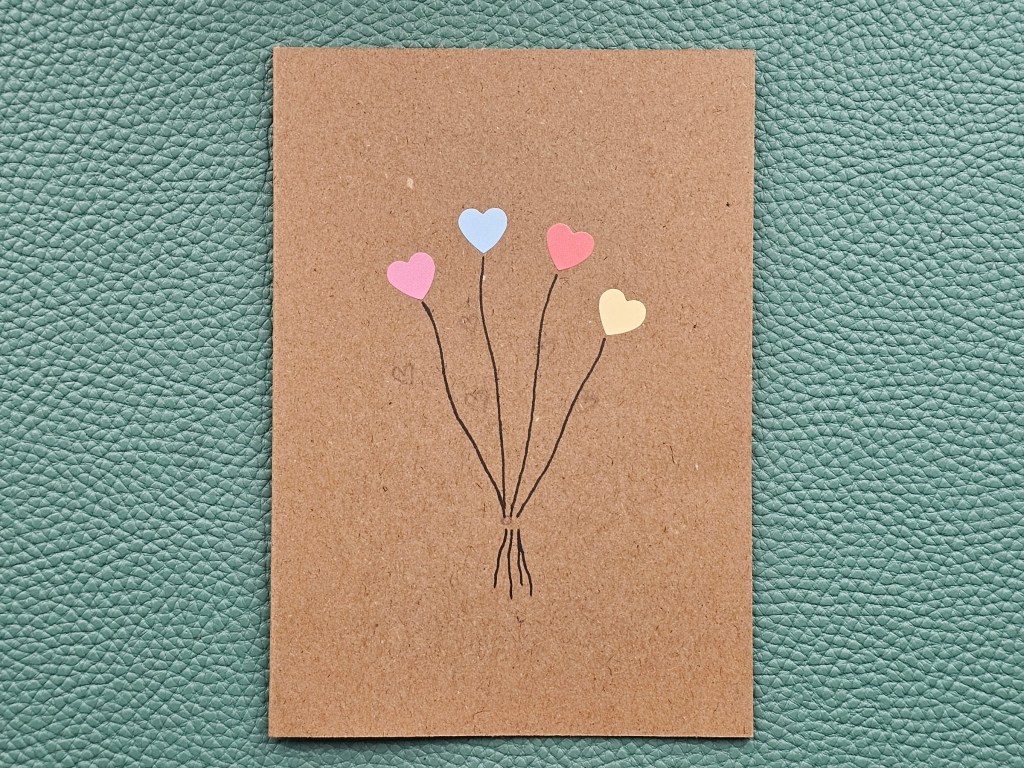 A brown paper card with four small heart shaped stickers arranged in an arch and strings drawn with black pen. A pencil sketch for the placement of other stickers can be seen on the card.