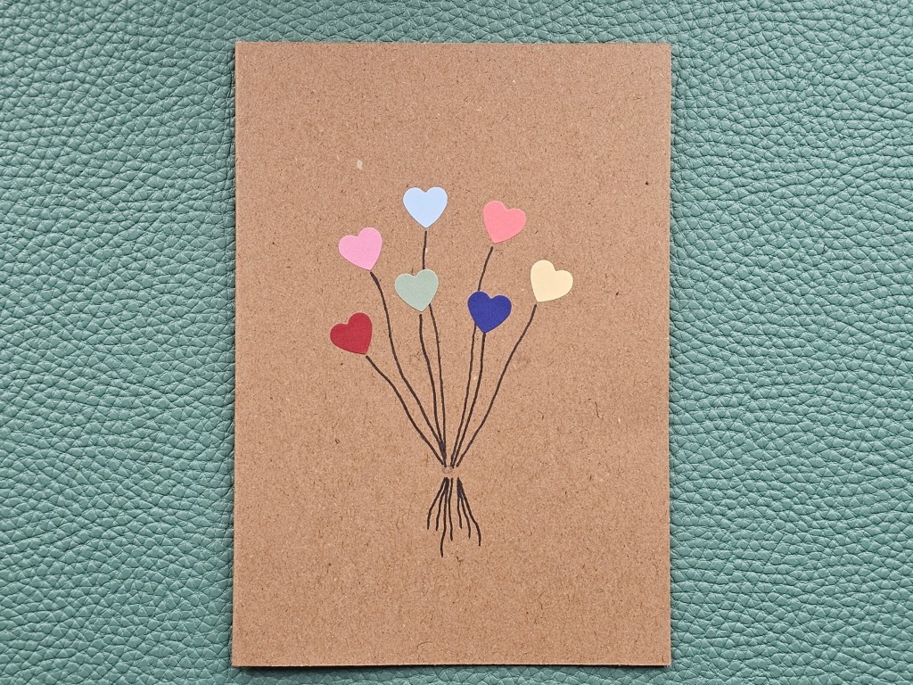 A brown paper card with seven small heart shaped stickers and strings drawn with black pen. A pencil sketch for the placement of other stickers can be seen on the card.