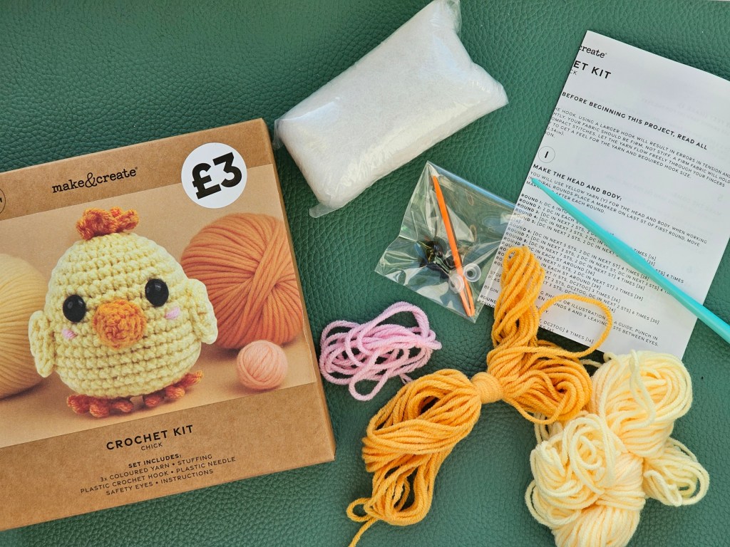 Contents of an assorted crochet set for crocheting a cute little chicken. It includes yarn in yellow, orange and pink, a crochet hook, toy stuffing, a plastic needle, safety eyes and instructions