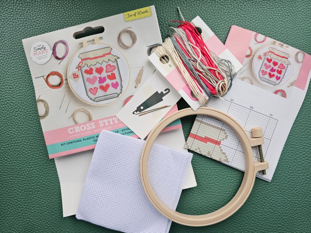 Contents of a cross stitch kit which includes a plastic embroidery hoop, a blank aida, threads in assorted colours, a needle, a threader, and a leaflet with instructions