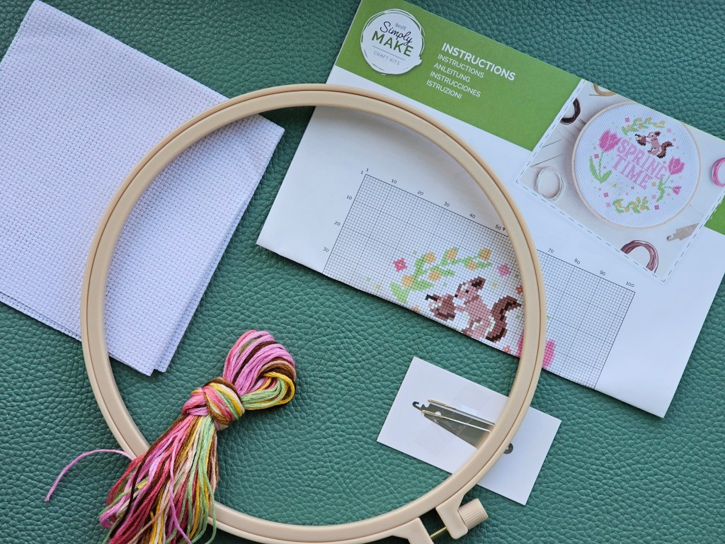 Contents of a cross stitch kit with a floral design including a squirrel. The set includes an embroidery hoop, a blank aida, assorted threads, a needle, a threader, and a leaflet with instructions.