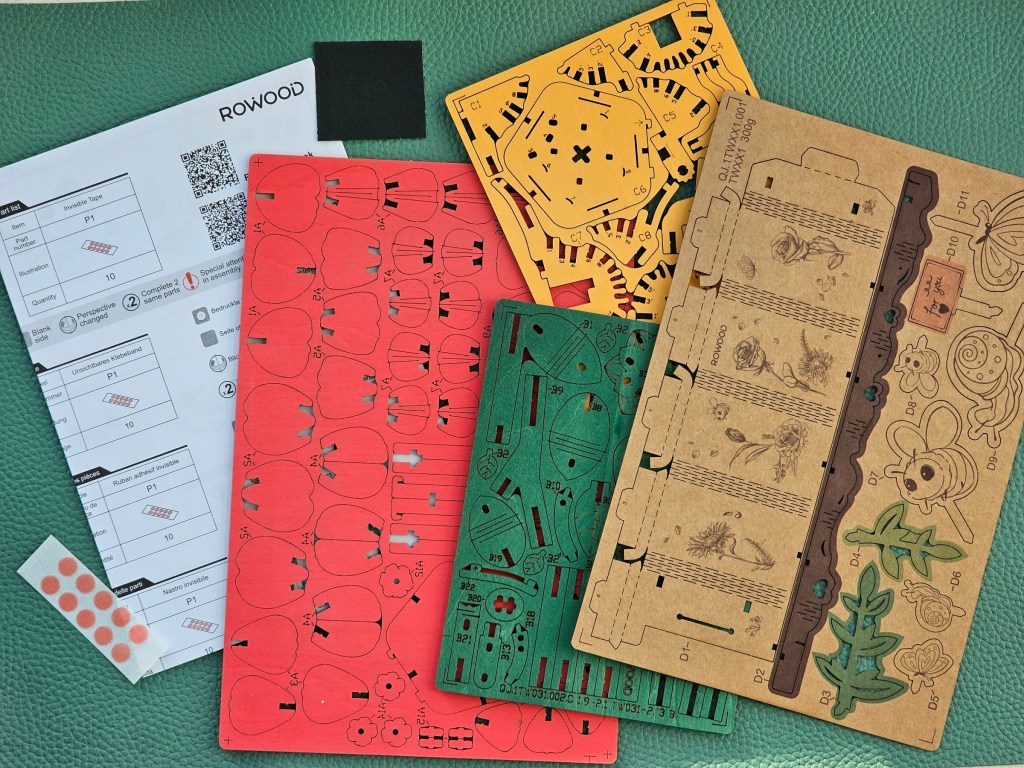 Contents of a set for building 3D wooden red camellia flowers. It includes pre-cut coloured wooden sheets, a leaflet with instructions, a piece of sandpaper and small pieces of adhesive double-sided tape