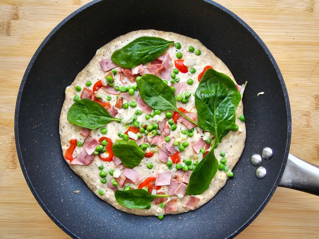 Cooked giant savoury pancake on a pan. There is ham, red pepper, green peas and spinach on the pancake.