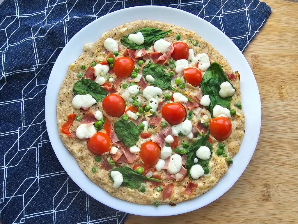 Cooked giant savoury pancake on a pan. There is ham, red pepper, green peas, spinach, cherry tomatoes and cheese on the pancake.