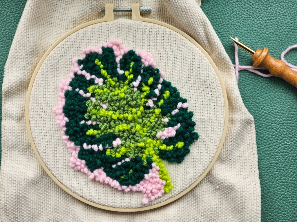 Nearly finished round punch needle canvas featuring a monstera leaf on a pink background