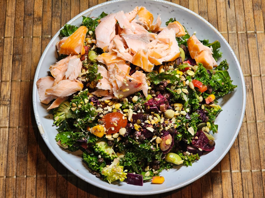 A plate with a generous serving of a salad made with kale, beetroot, tomatoes, corn, broccoli and lentils, served with hot smoked salmon