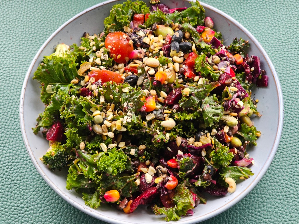 A plate with a generous serving of a salad made with kale, beetroot, tomatoes, corn, broccoli and lentils, sprinkled with seeds